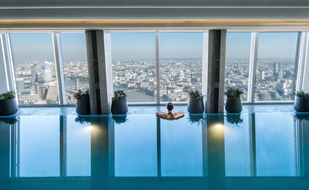 An indoor infinity-edge pool with one person seated at the far end faces floor-to-ceiling windows that look out over the London skyline and St Paul's Cathedral.
