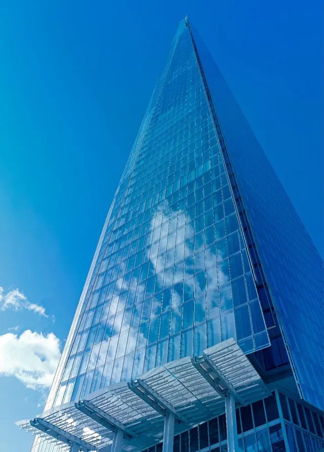 Looking up at The Shard, a tall glass-clad tower, against a clear blue sky, with a glass canopy at its base.