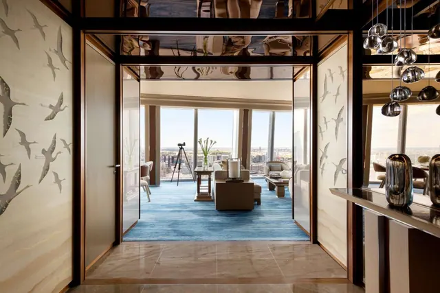 A suite entrance opens onto a living area with a blue carpet, seating, and floor-to-ceiling windows overlooking a city.