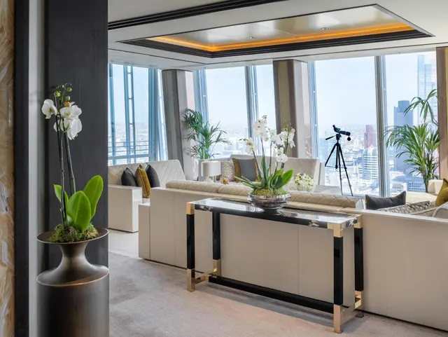 A living area with white sofas, a black and gold console table with white orchids, and floor-to-ceiling windows looking out over a city skyline.