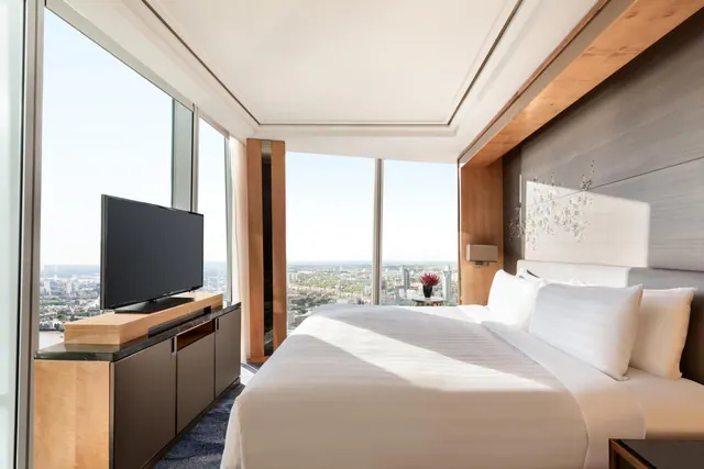 A bedroom with a made king bed, a television on a wood sideboard, and floor-to-ceiling windows overlooking a city.