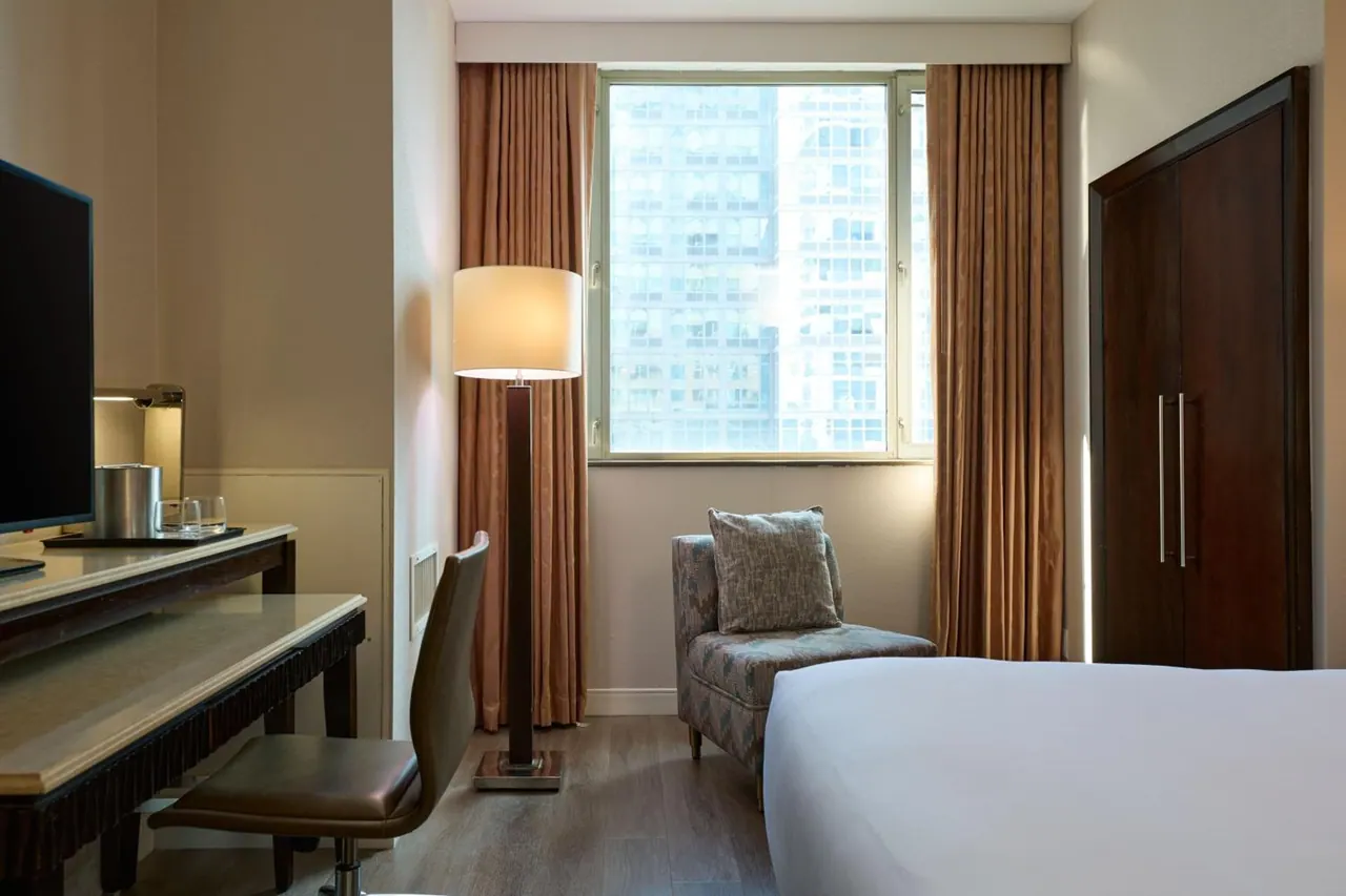 A hotel bedroom with a made bed, a desk and chair, a floor lamp, an upholstered armchair, a wooden wardrobe, and a window looking onto a high-rise building.