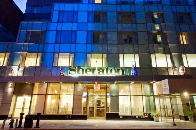 Glass-fronted hotel exterior at dusk with an illuminated Sheraton sign above the entrance, a canopy at the right, and flags on the facade.