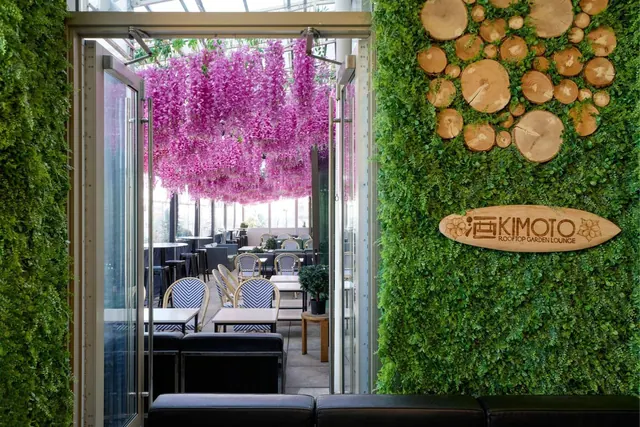 The entrance to the Kimoto Rooftop Garden Lounge, showing a living green wall, wood slice decor, hanging pink artificial wisteria, and tables with striped bistro chairs beyond the glass doors.