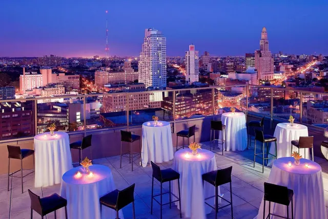 A rooftop terrace at dusk with white-clothed cocktail tables, lit candles and bar stools, overlooking a city skyline.