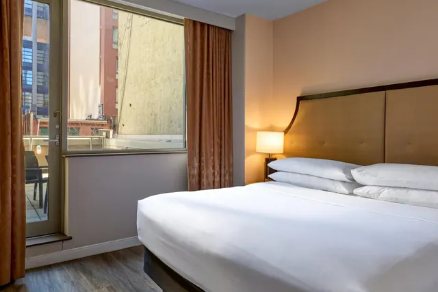 A made-up king bed with white pillows and an upholstered headboard sits beside a window and a glass door opening onto a balcony with chairs.