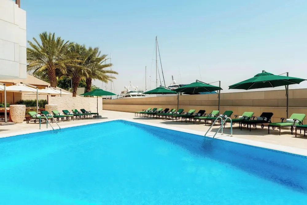 A rectangular outdoor swimming pool with green-cushioned sun loungers and green parasols, with yachts and palm trees beyond the wall.
