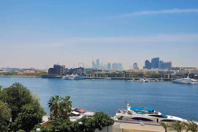 View across a wide waterway toward a city skyline with a tall tower, with moored yachts and palm trees in the foreground.