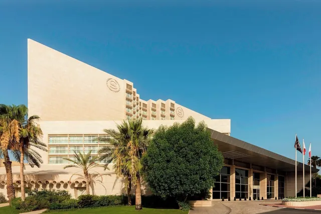 The exterior of a large cream-stone hotel building with a covered entrance, palm trees, flags and a large green tree under a clear blue sky.