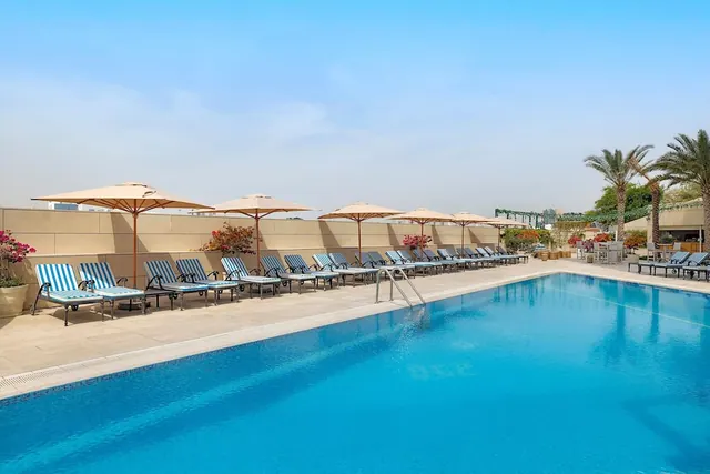A large outdoor swimming pool with striped sun loungers and beige umbrellas along the far edge, with palm trees to the right.