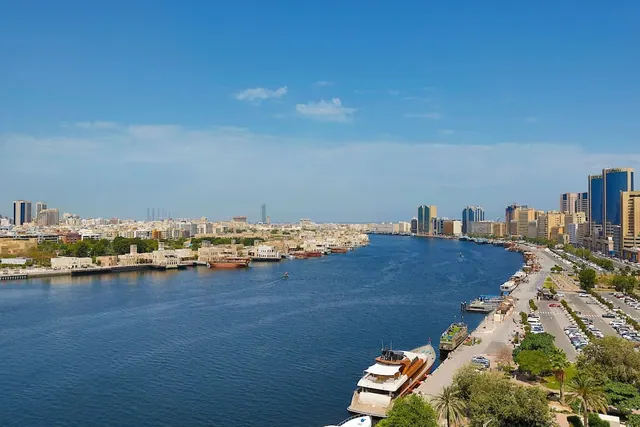 Elevated view over a wide waterway lined with low-rise buildings on the left, high-rise towers on the right, boats moored along the quay, and parked cars on a road.