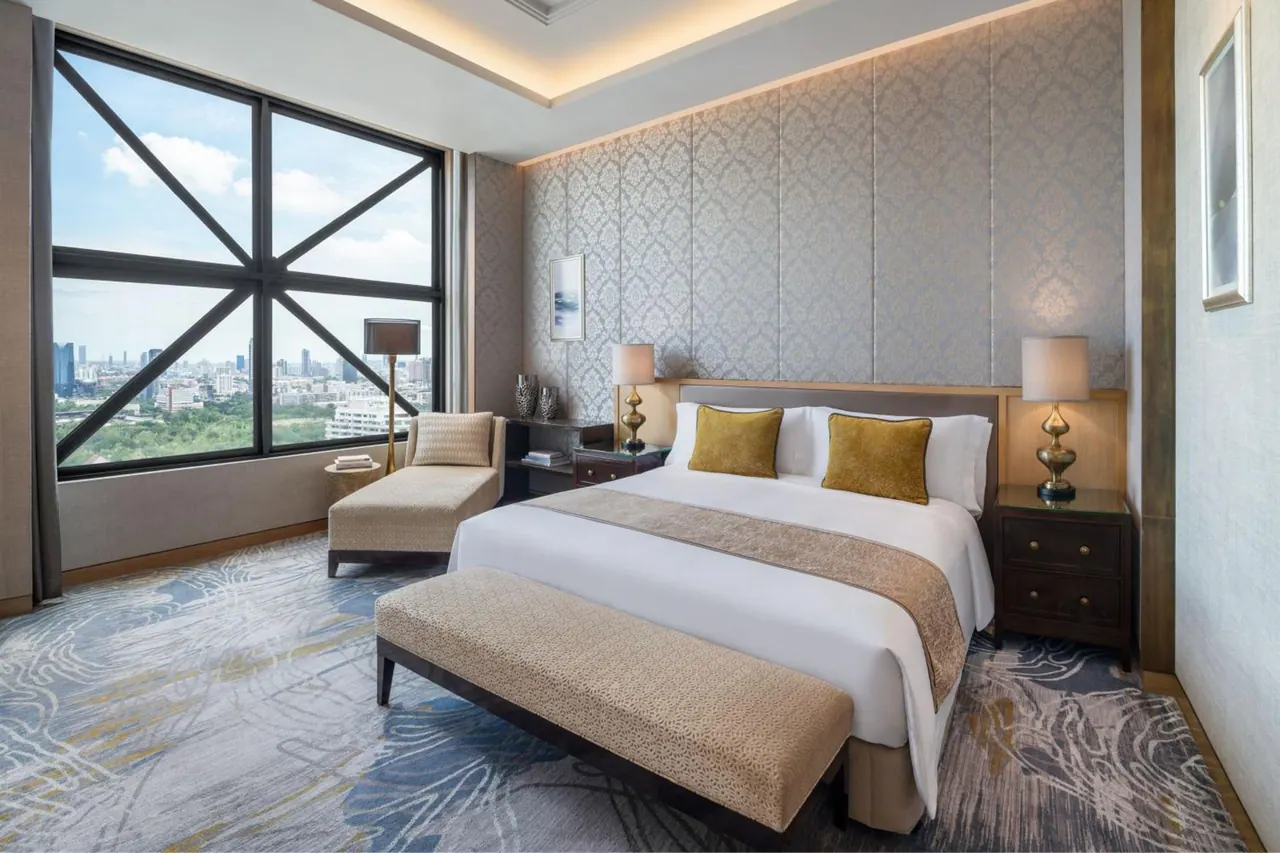 A bedroom with a king bed, a bench at its foot, and a lounge chair beside a large window overlooking a city skyline and green space.
