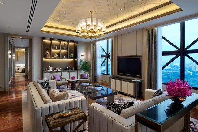 A staged living room with a sofa seating area, a wall-mounted television, a drinks cabinet and large windows looking out over a city skyline.