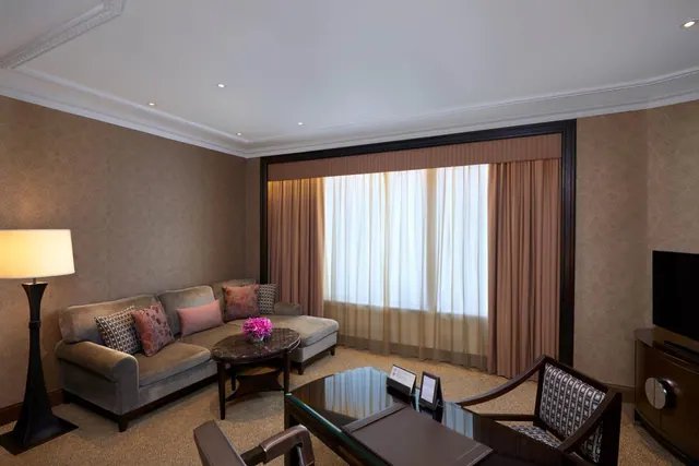 A hotel suite living area with a grey sofa, floor lamp, round side table with a pink flower arrangement, glass-topped desk with chair, and a wall-mounted television beside large sheer-curtained windows.