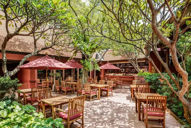 An outdoor dining terrace with wooden tables, red-cushioned chairs and red parasols under frangipani trees, with a food counter in the background.