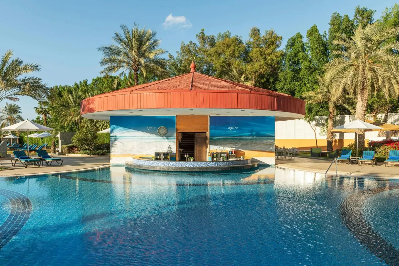 A large outdoor pool with a circular pool bar under a red roof, surrounded by palm trees, sun loungers and umbrellas.