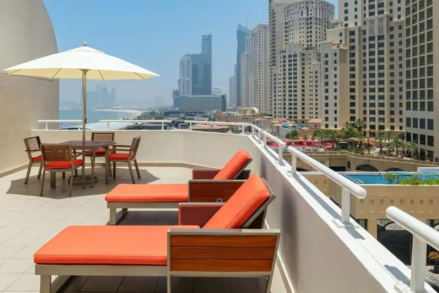 A furnished terrace with two orange sun loungers, a cream umbrella and a dining table overlooks high-rise towers, the sea and a pool below.