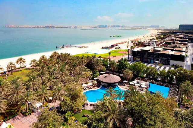 Elevated view of a resort with a swimming pool, palm trees, a sandy beach and the sea, with a city skyline in the distance.
