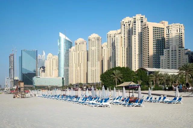 A sandy beach with rows of blue sun loungers and striped umbrellas, with high-rise towers rising behind it under a clear sky.