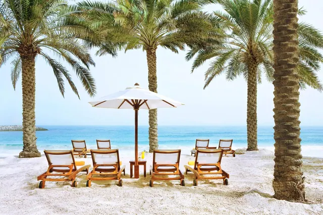 Wooden sun loungers and a white parasol stand on a sandy beach among palm trees, with the sea behind them.