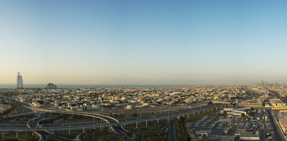 Aerial panoramic view over Dubai at dusk showing the Burj Al Arab, the sea in the distance, a multi-level highway interchange and a dense area of low-rise buildings.