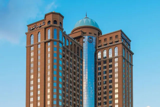 The upper section of a brown Sheraton hotel tower with a teal dome, a blue glass central column and Arabic and Latin signage against a clear blue sky.