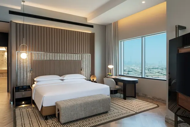 A bedroom with a king bed, an upholstered bench at its foot, a desk with a chair, and a wall-mounted television, with a window showing a hazy city view.