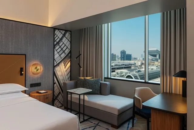 A hotel bedroom with a bed, a chaise lounge, a desk and a large window looking out over city buildings.
