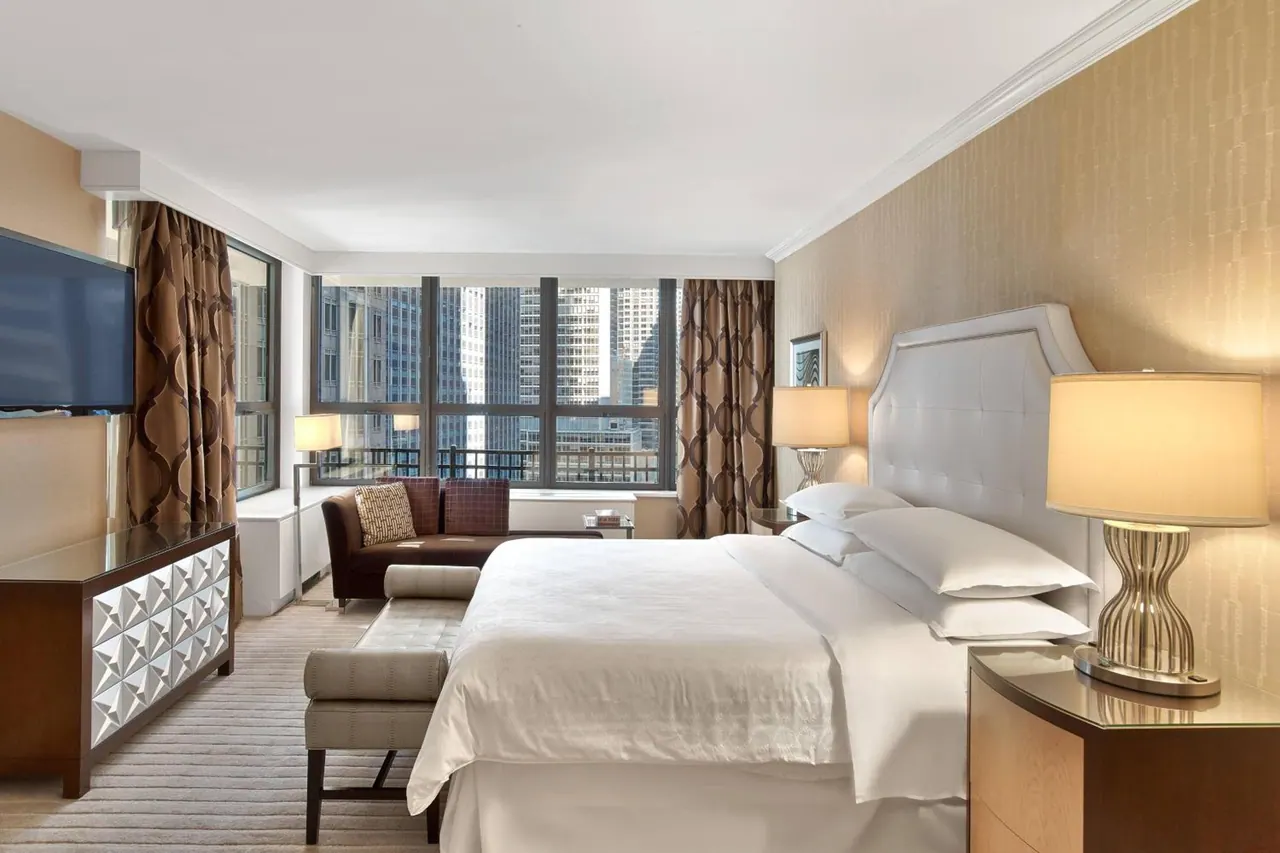 A hotel bedroom with a king bed, a brown sofa by floor-to-ceiling windows, a wall-mounted television, and a view of high-rise buildings outside.