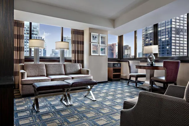 A living area with a grey sofa, two upholstered benches, a round table with chairs, and large windows showing city skyscrapers.