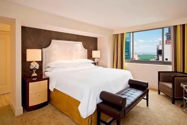 A bedroom with a king bed, a leather bench at the foot of the bed, an armchair, and a window overlooking city buildings and a green park.