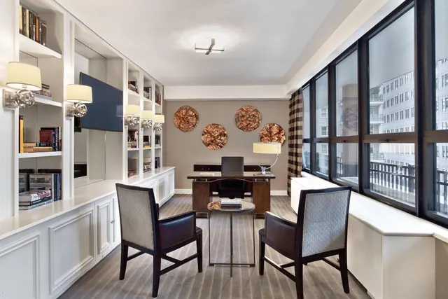 A study area with built-in white shelving, a wooden desk and chair, two upholstered armchairs and a round side table beside tall windows.