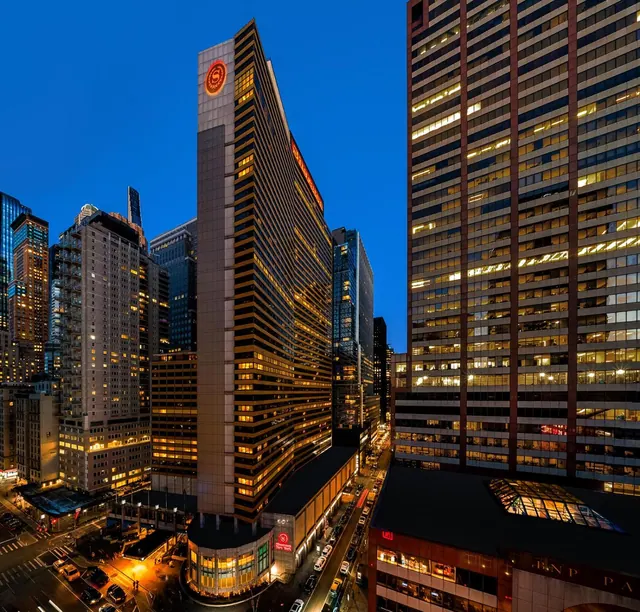 Twilight photo from above of a tall illuminated hotel tower with a red circular logo near the top, surrounded by office towers, with a street of traffic below.