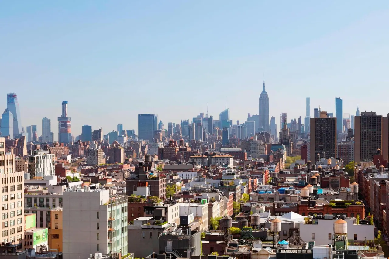 Elevated view over rooftops and low-rise buildings toward a Manhattan skyline that includes the Empire State Building under a clear blue sky.