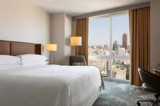 A king bed with white linens sits beside a floor-to-ceiling window overlooking a city skyline, with an armchair and a work desk nearby.