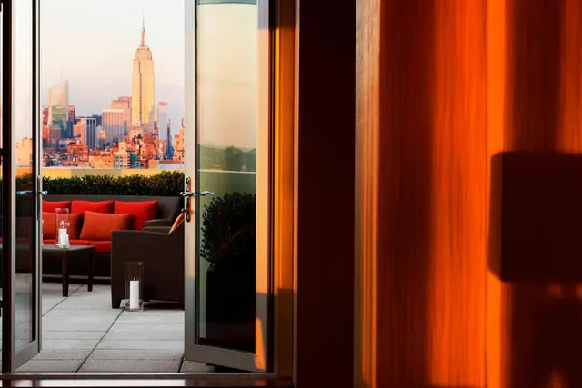 Open glass doors lead from a room onto a terrace with red cushioned wicker seating and a view of the Empire State Building.