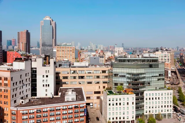 Elevated view over a dense cityscape of mid-rise and high-rise buildings, with water visible in the distance under a clear blue sky.