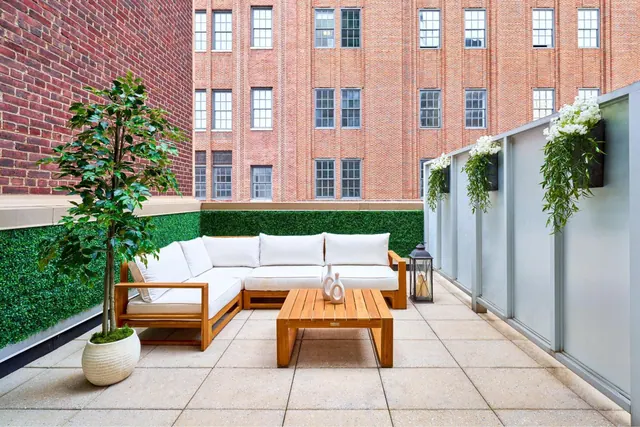 A furnished terrace with a white sectional sofa, a wooden coffee table and potted plants, with a brick building across the courtyard behind it.