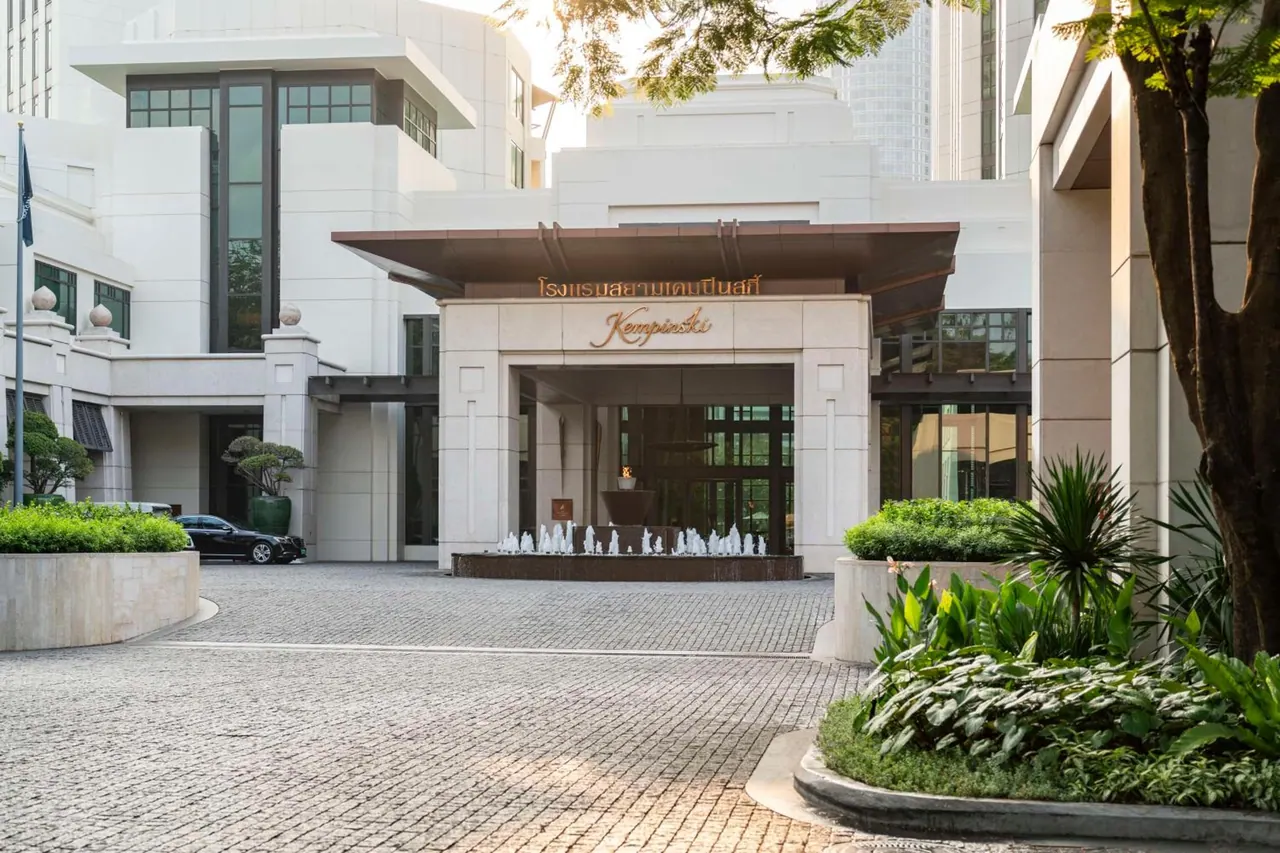 The hotel entrance has a covered drive with a fountain, a Kempinski sign above the doors, a cobbled driveway and a parked black car.