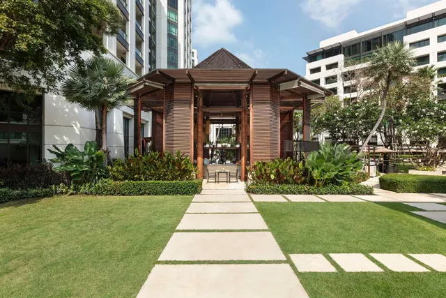 A stepping-stone path crosses a lawn to a wooden pavilion with a pitched roof, with tall buildings and palm trees around the garden.