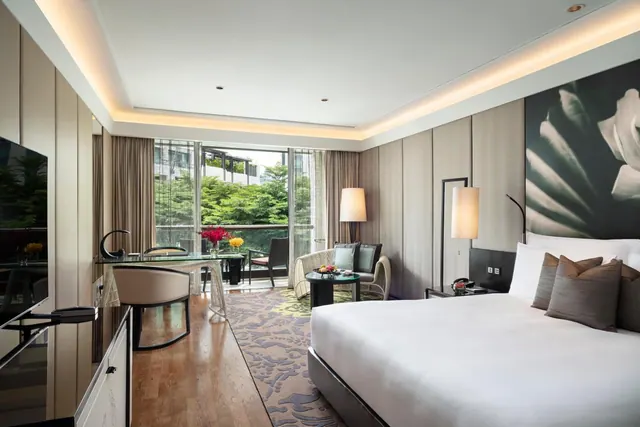 A hotel bedroom with a large bed, a seating area, a work desk and floor-to-ceiling glass doors opening onto a balcony with trees beyond.