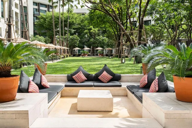 A sunken outdoor seating area with dark cushions and pink patterned pillows overlooks a lawn and tall trees, with potted cycads at the corners.