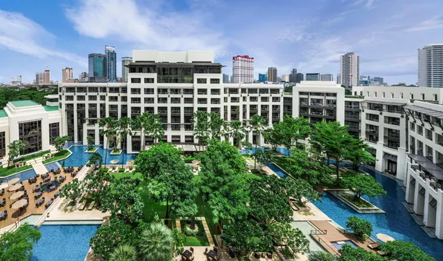 Aerial view of a white multi-storey hotel building around a landscaped courtyard with swimming pools, loungers, and a city skyline behind.