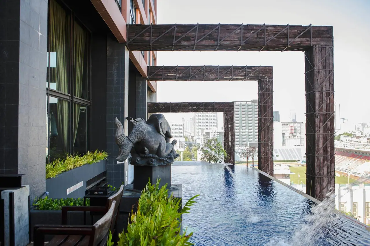 An infinity edge pool on a rooftop terrace with a bronze elephant sculpture, wooden pergola beams, and a view over city buildings and a stadium.