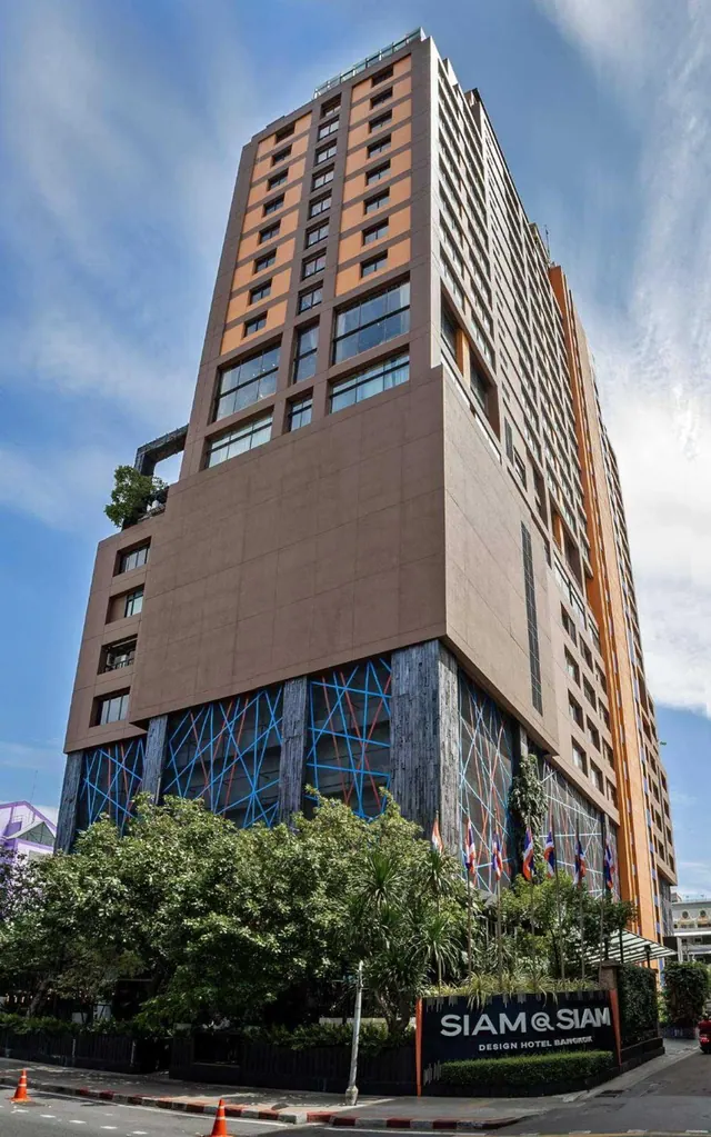 The exterior of the Siam@Siam Design Hotel tower is shown from street level under a blue sky, with trees and a traffic cone in front and a sign reading Siam@Siam at the entrance.