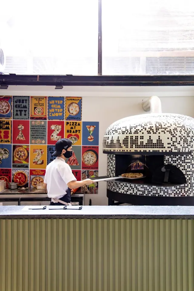 A cook in a cap and face mask lifts a pizza on a long peel from a mosaic-tiled wood-fired oven, with colourful pizza posters on the wall behind.