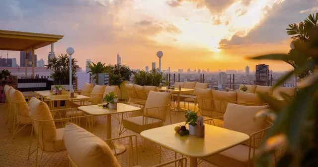 Outdoor rooftop seating area with yellow tables and cushioned chairs, overlooking a city skyline at sunset.