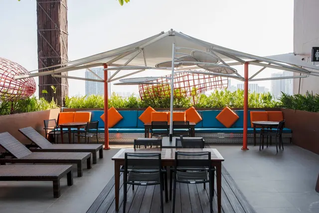 A rooftop terrace with an umbrella canopy, blue banquette seating with orange cushions, dining tables and sun loungers, with city towers visible in the hazy background.