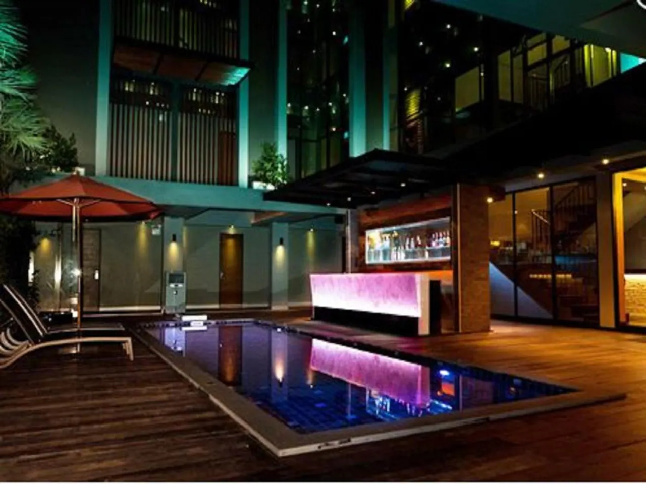 A small illuminated swimming pool on a wooden deck at night, with a purple-lit bar counter, sun loungers and a red umbrella beside it.