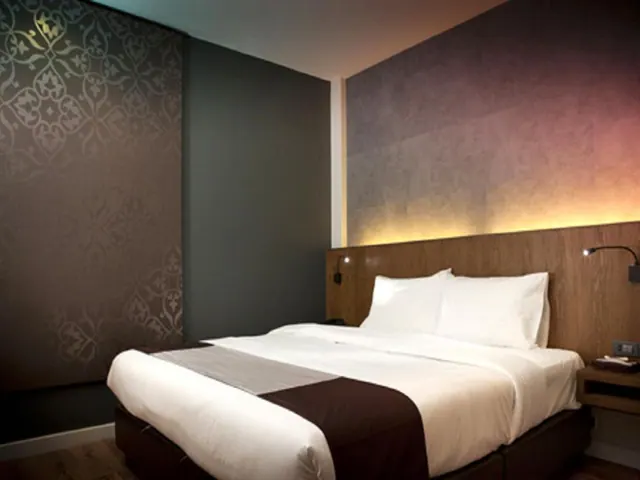 A double bed with white linens and a wooden headboard, lit by warm backlighting, sits beside a wall with patterned wallpaper.